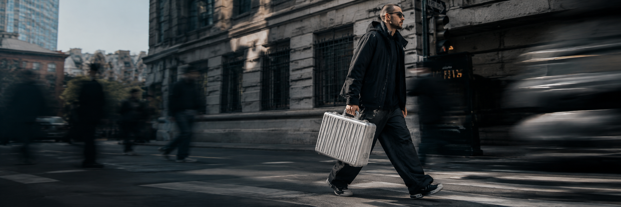 A man in dark clothing walks a city street holding a silver AC47 Case, cars and motion blur around him.